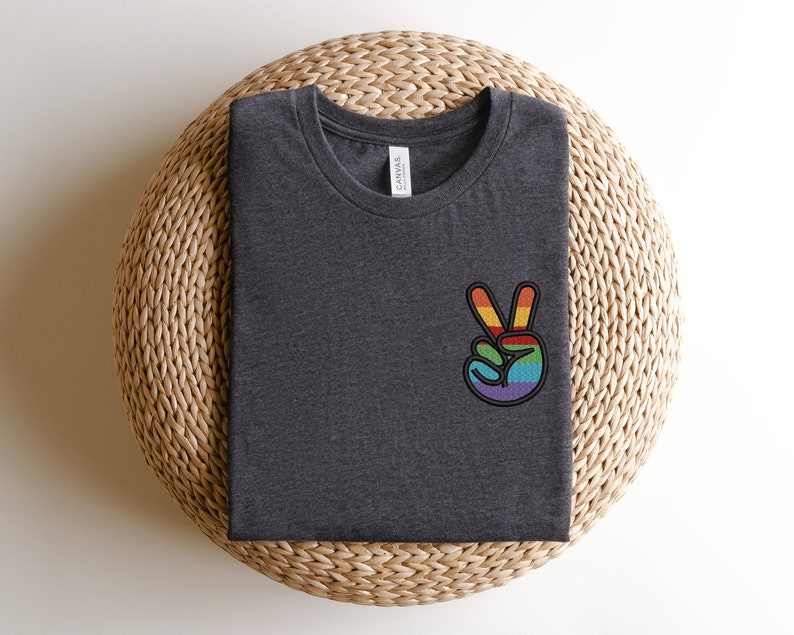 K&ouml;nnte beinhalten: Ein dunkelgraues T-Shirt mit einem gestickten Regenbogen-Friedenszeichen auf der linken Brustseite.