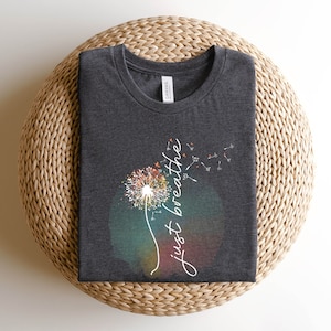 May include: Dark grey round-neck t-shirt with a dandelion design and the words "just breathe" in white script. The dandelion has a watercolour effect with orange, yellow, and green hues.