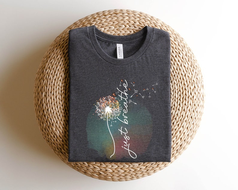 May include: Dark grey round-neck t-shirt with a dandelion design and the words "just breathe" in white script. The dandelion has a watercolour effect with orange, yellow, and green hues.