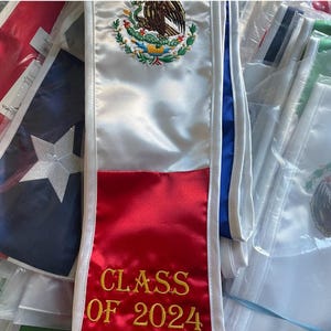 May include: A satin graduation stole featuring the Mexican coat of arms embroidered in the centre and the text CLASS OF 2024 stitched in gold thread on the red section. The stole is white with a red bottom panel and blue trim along the edges.