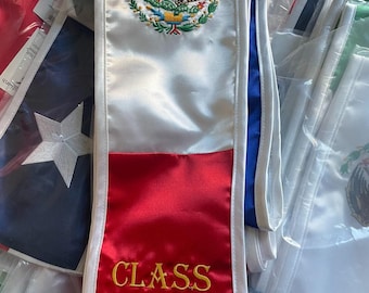 May include: A satin graduation stole featuring the Mexican coat of arms embroidered in the center and the text CLASS OF 2024 stitched in gold thread on the red section. The stole is white with a red bottom panel and blue trim along the edges.