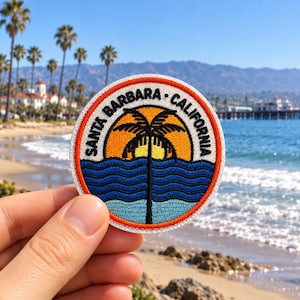 May include: A circular embroidered patch featuring a black palm tree silhouette against a yellow and orange sun, set above blue wavy ocean water. The text SANTA BARBARA CALIFORNIA is stitched in black along the top edge, bordered by an orange rim.
