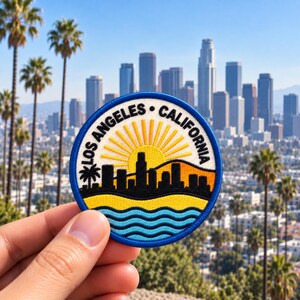 May include: A circular embroidered patch featuring a black city skyline silhouette against a yellow sunburst. The text LOS ANGELES CALIFORNIA is arched across the top. Below the skyline are wavy blue lines representing water, all bordered by blue.