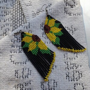 May include: A pair of dangle earrings featuring black, yellow and green beads. The beads are arranged in a pattern resembling a sunflower. The earrings are made with silver wire.