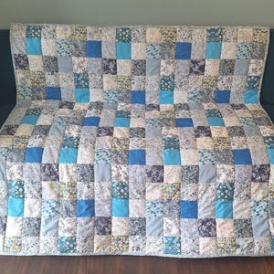 May include: A rectangular patchwork quilt featuring a grid of squares in various shades of blue, white, and grey. The design incorporates solid blue squares interspersed with squares displaying floral, botanical, and geometric patterns throughout.
