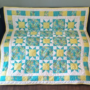 May include: A rectangular quilt featuring a repeating pattern of star blocks and floral print octagons. The design incorporates bright yellow, turquoise, and white fabric squares with a yellow border trim, creating a vibrant, geometric textile.