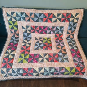 May include: A colourful quilt with a pinwheel design. The quilt features squares of various colours, including pink, blue, green, and red. The quilt is bordered with a floral pattern and a dark blue trim.