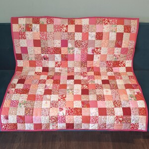 May include: A patchwork quilt featuring a grid of square fabric patches in various shades of pink, red, and white. The squares display diverse floral, polka dot, and geometric patterns. The quilt is finished with a pink border with white dots.