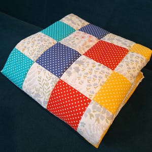 May include: A colourful patchwork quilt with squares in various patterns. The squares feature polka dots and floral prints in shades of blue, red, yellow, and white. The quilt is folded, appearing soft and comfortable, and is likely made of cotton.