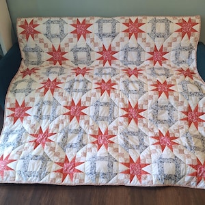 May include: A rectangular quilt featuring a repeating pattern of eight-pointed stars and square blocks. The design incorporates red floral fabric, blue and white patterned fabric, and solid white fabric, all stitched together in a grid layout.