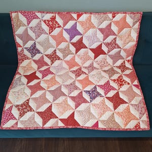 May include: A rectangular patchwork quilt featuring a repeating star pattern composed of various fabric squares in shades of red, pink, and white. The design incorporates floral, polka dot, and solid prints, all bordered by a solid pink fabric trim.