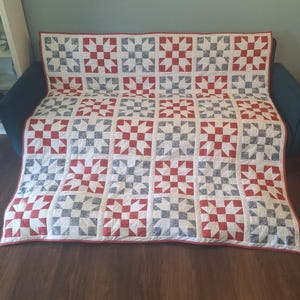 May include: A rectangular quilt featuring a repeating block pattern in red, white, and dark blue. The design consists of alternating star and grid motifs constructed from fabric squares. The entire quilt is finished with a thin red border.
