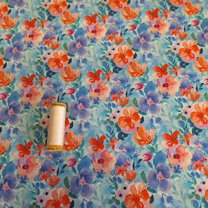 May include: A light blue fabric featuring a repeating floral pattern of orange and purple watercolour-style flowers with green leaves, accompanied by a small spool of light-coloured sewing thread placed on the surface of the patterned textile.