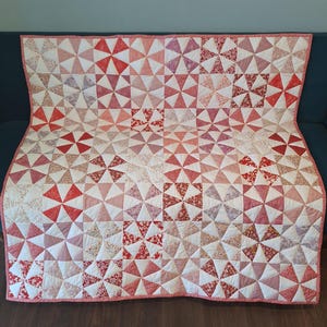 May include: A rectangular quilt featuring a repeating pinwheel block pattern. The design consists of various shades of red, pink, and white fabric, with some blocks displaying floral prints and others solid colours, finished with a thin red border.