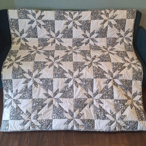 May include: A handmade quilt with a starburst pattern. The quilt features alternating white squares and squares with a dark blue floral print. The quilt is folded over a dark blue sofa.