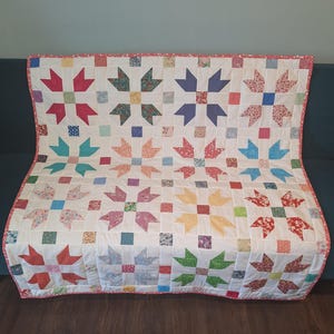May include: A rectangular patchwork quilt featuring a repeating star block pattern. The quilt is constructed with white fabric squares and various multi-coloured floral and geometric patterned fabric pieces, finished with a thin pink border.