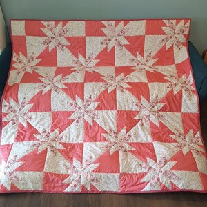 May include: A rectangular quilt featuring a repeating star pattern. The design alternates between solid coral pink fabric with a subtle white dot pattern and a white fabric printed with a delicate pink and green floral motif throughout.