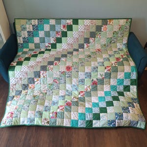 May include: A patchwork quilt featuring a grid of square fabric patches in various shades of green, white, and teal. The design incorporates solid colours alongside floral and botanical patterns, finished with a solid dark green fabric border.