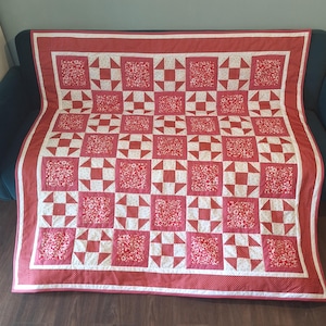 May include: A rectangular quilt featuring a repeating pattern of red and white floral squares alternating with red and white geometric blocks. The quilt is framed by a wide red border with a white dotted pattern and a thin white inner border.