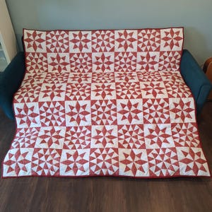 May include: A rectangular quilt featuring a repeating pattern of red and white eight-pointed stars. The design consists of alternating blocks of star motifs and solid white squares, finished with a thin red border along the outer edges.