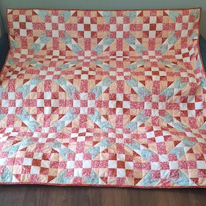 May include: A rectangular quilt featuring a repeating geometric pattern of nine-patch blocks and triangles. The fabric displays a colour palette of soft pink, coral, white, and light blue, with small floral and dotted prints throughout the design.