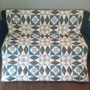 May include: A rectangular quilt featuring a geometric pattern of diamonds and squares. The fabric displays a mix of white, teal, and dark green floral prints, with small red accents throughout the design. The quilt is stitched with a grid layout.