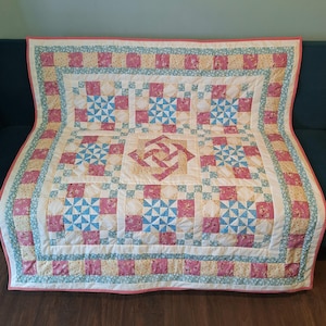 May include: A handmade quilt with a patchwork design. The quilt features a central square with a pink and white pinwheel pattern, surrounded by squares of pink, blue, and floral fabrics. The border is a mix of floral and striped patterns, with a red and white striped edge.