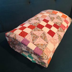 May include: Folded patchwork quilt with red, white, pink, and floral fabrics. The quilt has a checkerboard design with squares and triangles. It is neatly folded on a dark teal surface. The quilt is made of various fabrics.
