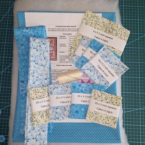 May include: A quilt kit containing fabric pieces, batting, and a spool of thread. The kit includes labelled bundles of blue and cream floral fabric squares, blue polka dot fabric, and an instruction sheet titled Cosysews Nine Stars Quilt Kit.