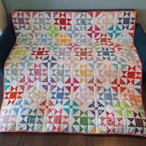 May include: A colourful patchwork quilt with star patterns in shades of red, blue, yellow, and green. The quilt is made from fabric squares and has a white background with a pink border. It is draped over a dark blue sofa, creating a cozy aesthetic.