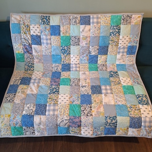 May include: A patchwork quilt in shades of blue, white, and teal. The quilt features square patches with floral, polka dot, and gingham designs. It is bordered with a light grey fabric and would suit a cot or small bed.