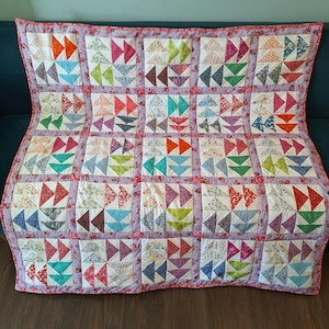 May include: A patchwork quilt featuring a repeating pattern of colourful triangles in shades of red, blue, green, and orange. The quilt has a floral border and is displayed on a dark blue surface. The quilt is a handmade item.