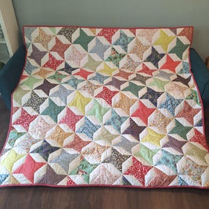 May include: A rectangular quilt featuring a repeating star pattern composed of various floral and solid-coloured fabric patches in shades of red, yellow, green, blue, and white. The quilt is finished with a solid pink fabric border trim.