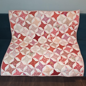 May include: A rectangular quilt featuring a repeating star pattern composed of various fabric squares in shades of red, pink, and white. The design includes floral, polka dot, and solid prints arranged in a grid, finished with a thin patterned border.
