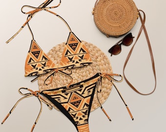Op de afbeelding: Een bruine en zwarte bikini met een driehoekige top en een hoge snit. De bikini ligt op een strooien mat met een strooien tas en een zonnebril.