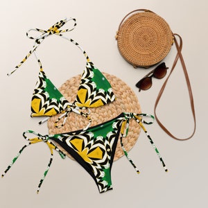 May include: A green, yellow and black patterned bikini with a matching straw bag and sunglasses. The bikini top is a triangle style with adjustable straps. The bikini bottom is a cheeky style with ties on the sides.