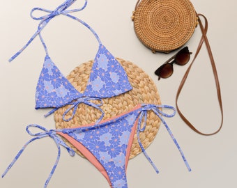 May include: A blue and pink floral print bikini with a matching top and bottom. The bikini is on a woven straw mat with a straw bag and sunglasses.
