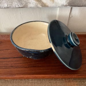 May include: A small, round ceramic bowl with a lid. The bowl is dark blue with a cream-colored interior. The lid is slightly ajar, revealing the inside of the bowl. The bowl is sitting on a wooden surface.