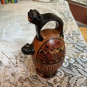 May include: A terracotta-colored ceramic vessel featuring a black feline-shaped handle and a smaller black feline figure protruding from the side. The body is decorated with intricate geometric patterns, red and black accents, and a small llama motif.