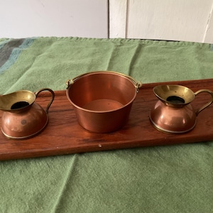 May include: Three copper and brass decorative items on a wooden tray. The items include two small pitchers and a small bucket with a handle. The pitchers have a brass top and handle. The bucket is round with a small handle.