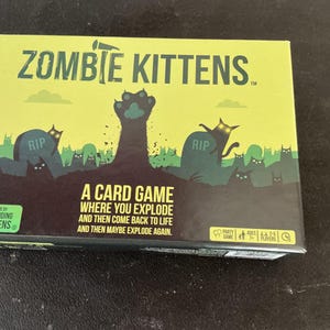 May include: A box for the card game "Zombie Kittens" with a lime green background. The box features a graveyard scene with zombie cats and the text "Zombie Kittens". The game is by Exploding Kittens.