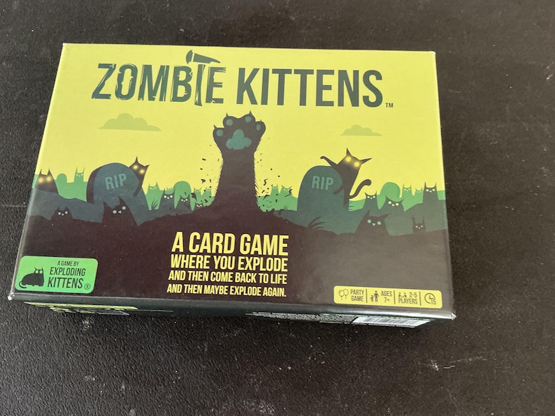 Może przedstawiać: Pudełko do gry karcianej "Zombie Kittens" z limonkowym tłem. Pudełko przedstawia scenę cmentarza z kotami zombie i tekstem "Zombie Kittens". Gra jest autorstwa Exploding Kittens.
