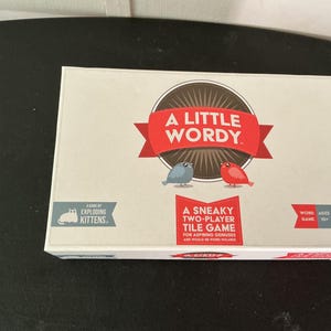 May include: A Little Wordy board game box. The box is white with red and brown accents. The game is by Exploding Kittens. The box features the text "A Little Wordy" and "A Sneaky Two-Player Tile Game".