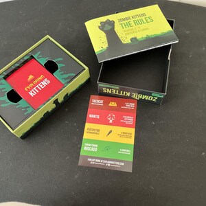 Może przedstawiać: Otwarty zestaw pudełkowy gry karcianej "Exploding Kittens" z czerwonym pakietem kart w środku. Pudełko jest zielono-czarne z napisem "Exploding Kittens" i "Zombie Kittens". Widoczna jest r&oacute;wnież oddzielna książeczka i karta.