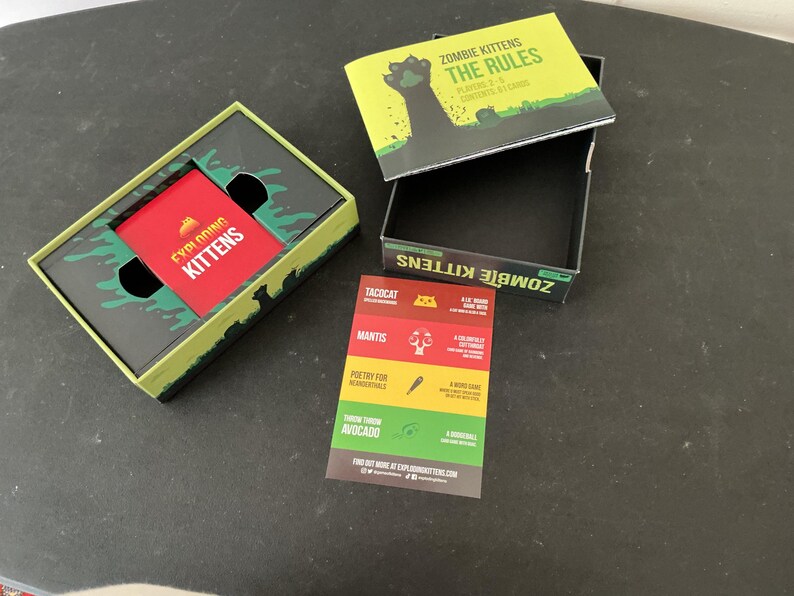 Może przedstawiać: Otwarty zestaw pudełkowy gry karcianej "Exploding Kittens" z czerwonym pakietem kart w środku. Pudełko jest zielono-czarne z napisem "Exploding Kittens" i "Zombie Kittens". Widoczna jest r&oacute;wnież oddzielna książeczka i karta.