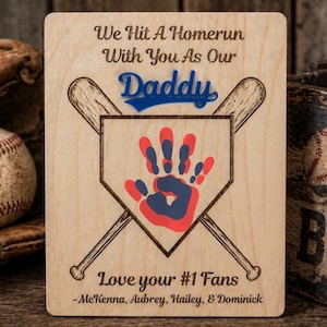 May include: Wooden plaque with baseball theme. Features crossed bats, a home plate with a red and blue handprint, and the words "We Hit A Homerun With You As Our Daddy." Includes the text "Love your #1 Fans."