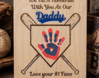 Puede incluir: Placa de madera con temática de béisbol. Presenta bates cruzados, un plato de home con una huella de mano roja y azul, y las palabras "We Hit A Homerun With You As Our Daddy". Incluye el texto "Love your #1 Fans."