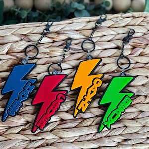 May include: Four keychains in the shape of lightning bolts, each with a different color: blue, red, orange, and green. Each keychain has a black chain and ring. The text on each keychain is in a matching color.