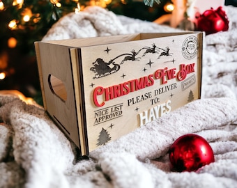 May include: A wooden Christmas Eve box with laser-cut Santa and reindeer designs. The box has the words "CHRISTMAS EVE BOX" in red and "PLEASE DELIVER TO: HAYES" in white. A red Christmas ornament sits nearby on a soft, white blanket.