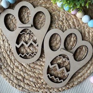 May include: Two wooden Easter egg holders shaped like paw prints. Each paw print has three egg-shaped cutouts and a zig-zag patterned egg in the center.